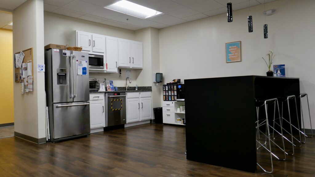 Image of an office breakroom with seating and a fridge, sink, coffee bar with wall art and nice lights.