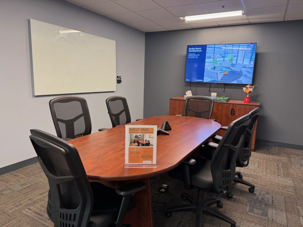 A small meeting room with no windows for six to eight people. Room has a white board and a tv screen, and some mesh backed chairs.