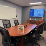 A small meeting room with no windows for six to eight people. Room has a white board and a tv screen, and some mesh backed chairs.