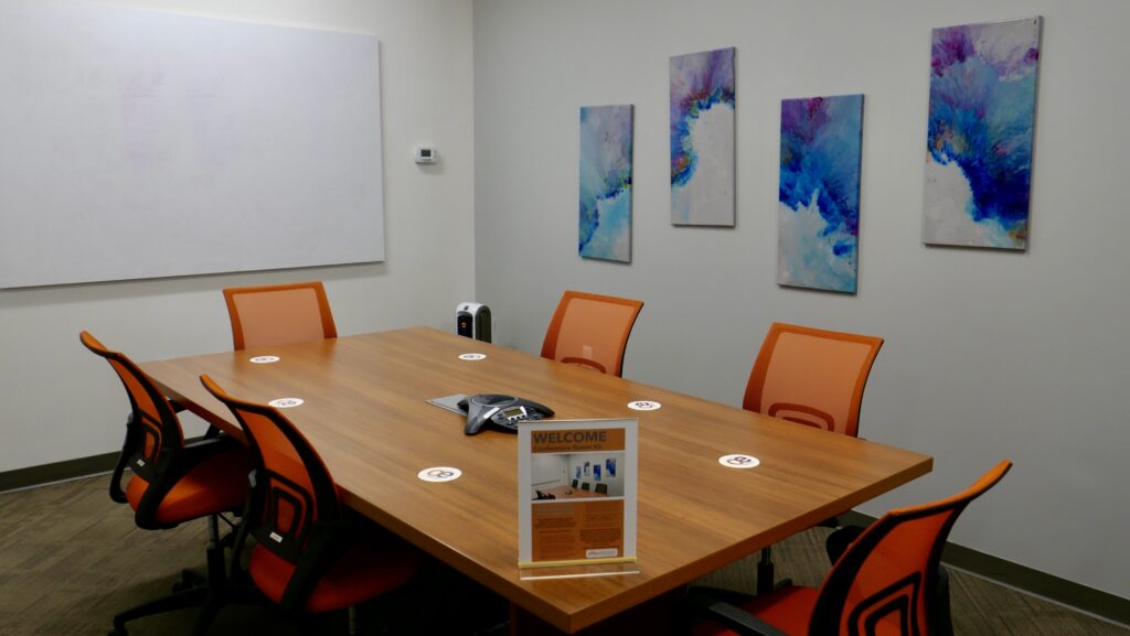 A six person meeting room with orange chairs and a large white board on the wall along with some wall mounted paintings.