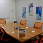 A six person meeting room with orange chairs and a large white board on the wall along with some wall mounted paintings.