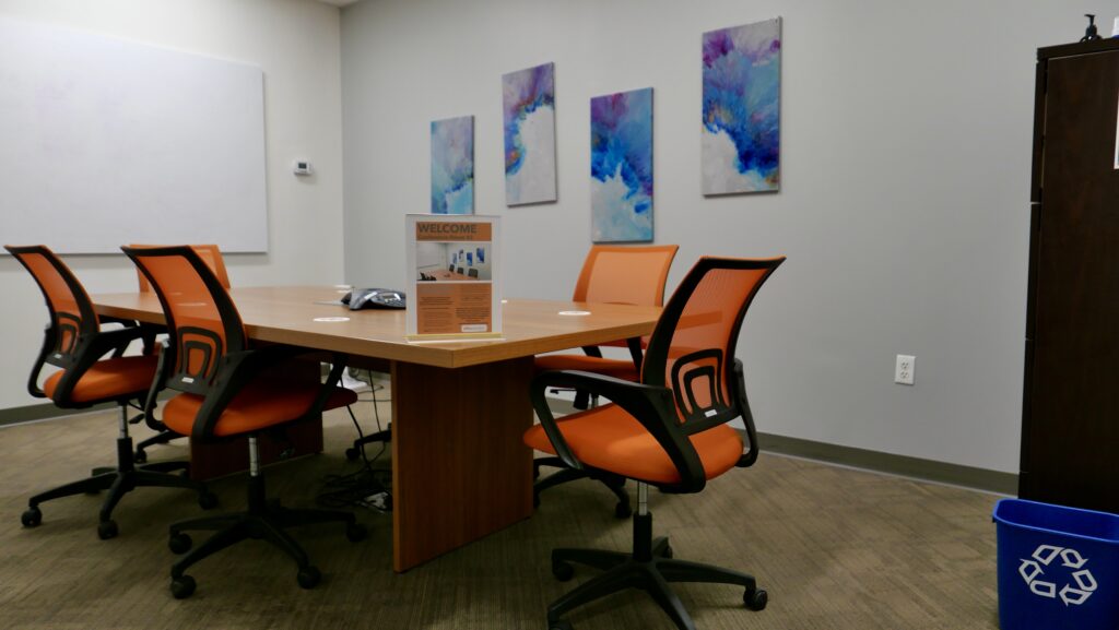 A medium sized meeting room space with colorful artwork on the wall and orange colored chairs.