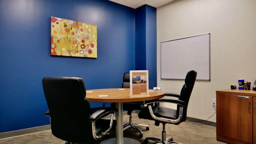 A small meeting room with no windows but a nice painting and a wall mounted white board. Blue walls. Seating for four people.