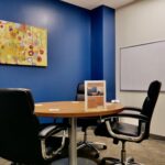 A small meeting room with no windows but a nice painting and a wall mounted white board. Blue walls. Seating for four people.