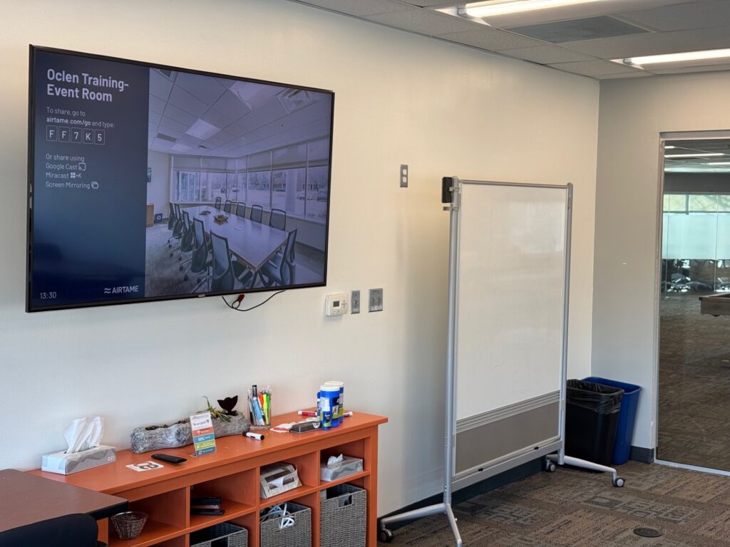 Image of a sizeable television flat screen and a wheel-mounted white board in a training room.