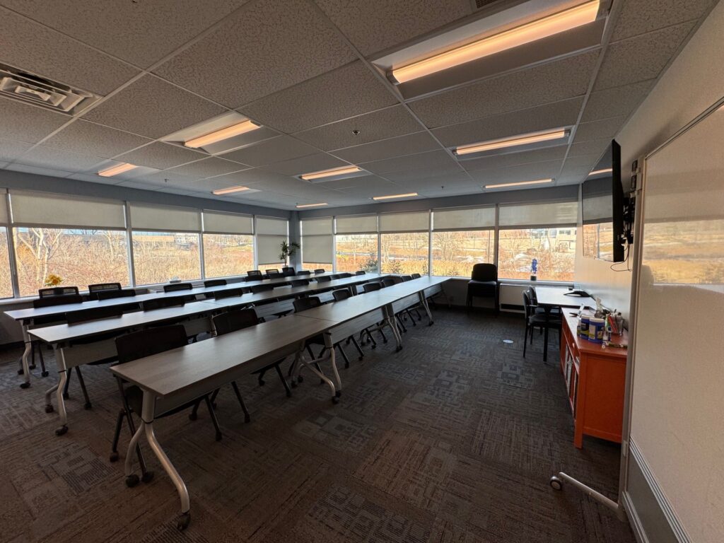 A large modern office training room space with seating for 30 people on rolling chairs. Room has sunny windows on two sides.