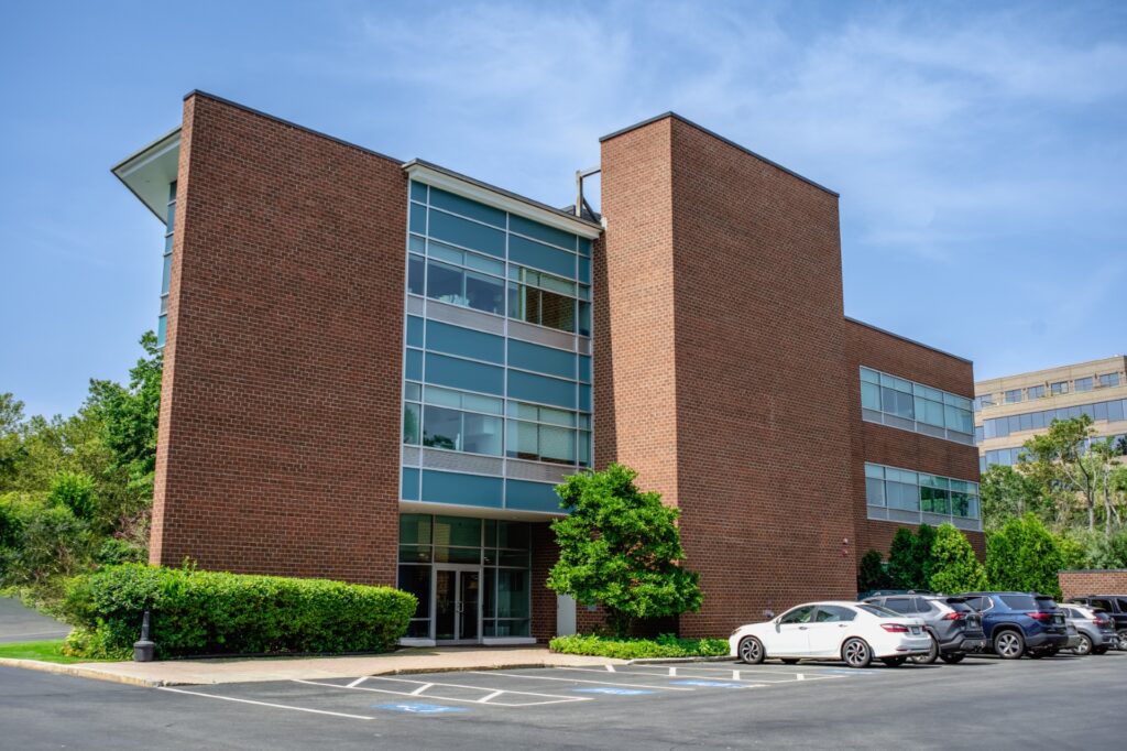 A tall brick office building with green trees and cars parked outside.