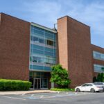 A tall brick office building with green trees and cars parked outside.